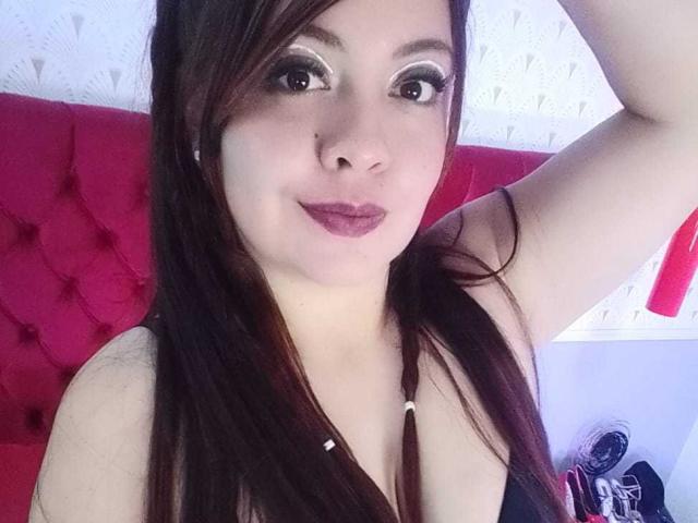 AmatisthaBlend has dark brown hair, wearing a black top, showing her tits with a raised arm, and has purple lipstick, with a red tufted headboard in the background.