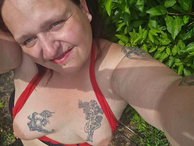 Hotsunny20-sex, red bikini top, fair-skinned, tattoos on chest, nipple piercings, nose ring, smiling, green leaf background, brown hair, arm tattoos, selfie, outdoors, sunlit.