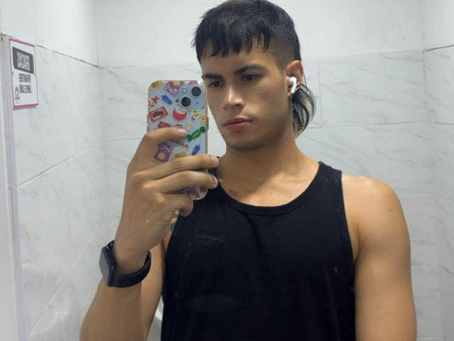 DallasSi, con cabello negro corto, audífonos blancos, una camiseta negra sin mangas y un reloj de pulsera negro, sosteniendo un teléfono de colores, tomando una selfie frente al espejo en un baño con azulejos blancos..