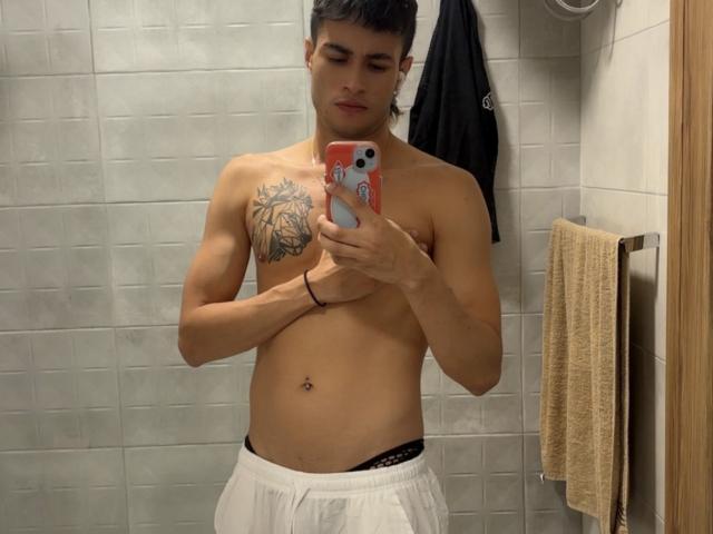 Dallas, senza camicia, che tiene un telefono, con un tatuaggio nero sul petto, pantaloni bianchi, biancheria intima nera, piercing all'ombelico, capelli corti e scuri, selfie allo specchio, espressione neutra, bagno, asciugamano a destra..
