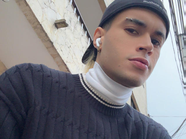 DallasSi, con una expresión seria, lleva puesto un gorro negro, un suéter negro de punto acanalado, un cuello alto blanco y auriculares blancos. Tiene un piercing en la nariz y auriculares en el pelo..