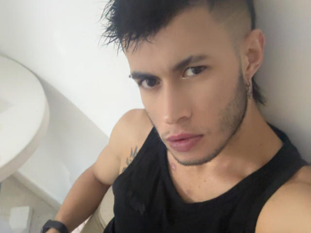 DallasSi has a muscular, tan-skinned face with short, spiked black hair, brown eyes, and a small tattoo on his neck. He's wearing a black tank top, looking serious.