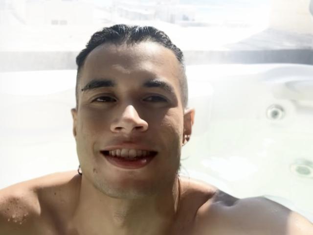 DallasSi, zonder shirt, in een jacuzzi met donker, nat haar, kleine zwarte oorbellen en een ontspannen glimlach, met licht vuil water op zijn gezicht en borst..