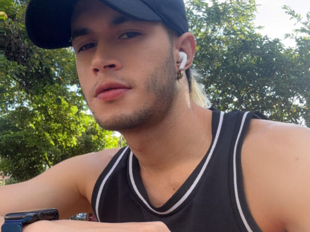DallasSi mit leichtem Bart, schwarzer Kappe, weißen Kopfhörern, schwarzem Tanktop und einer Uhr, entspannt im Freien, mit grünen Bäumen im Hintergrund..