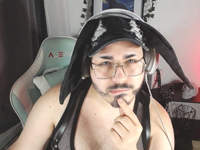 FabianNoLimits with glasses, black headband, and headphones, chest hair, finger on chin, tank top, sitting in a gaming chair. Bearded, slightly chubby, looking at camera.