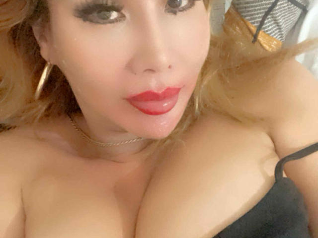 QueenFhukelya mit rotem Lippenstift, schwarzem Träger und großen Brüsten, trägt goldene Ohrringe und eine goldene Kette, liegt, hat blondes Haar und ein leicht glänzendes Gesicht..
