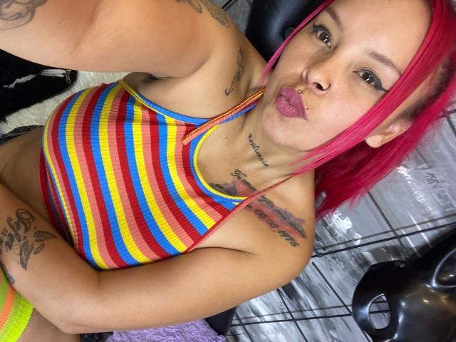 AlanaRoth with pink hair, pouty lips, striped yellow-red-blue tank top, tattoos on arm and chest, holding game controller, looking at camera, showing her small tits.