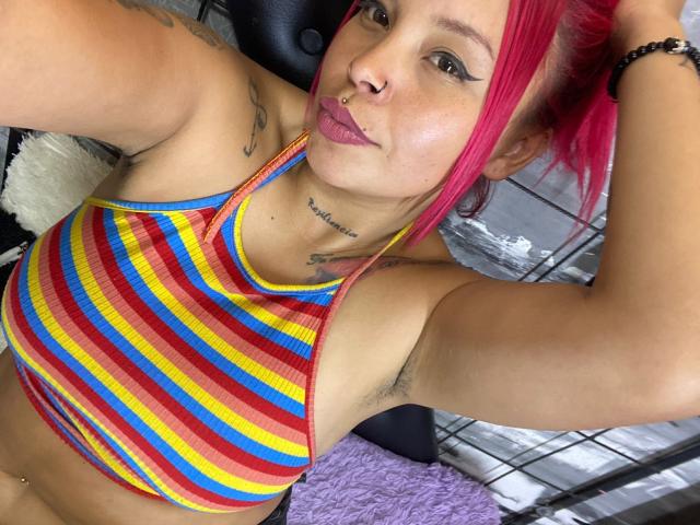 AlanaRoth with pink hair, striped yellow-red-blue top, nose ring, tattoos, and pouty lips, showing armpit hair and small tattoos, taking a selfie. Her boobs are visible.