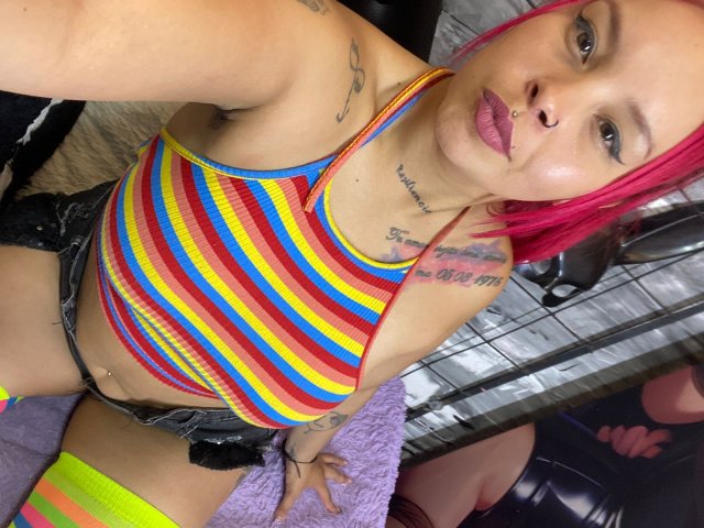 AlanaRoth with pink hair, striped yellow-red-blue top, black shorts, neon green knee socks, tattoos, nose ring, and lip ring, showing her tits and pussy in a selfie.