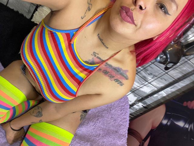 Alana Roth, con cabello rosa, piercing en la nariz y tatuajes, vestida con un top corto a rayas de arcoíris y medias altas a rayas verde neón y rosa, mostrando sus pequeños pechos y glúteos..