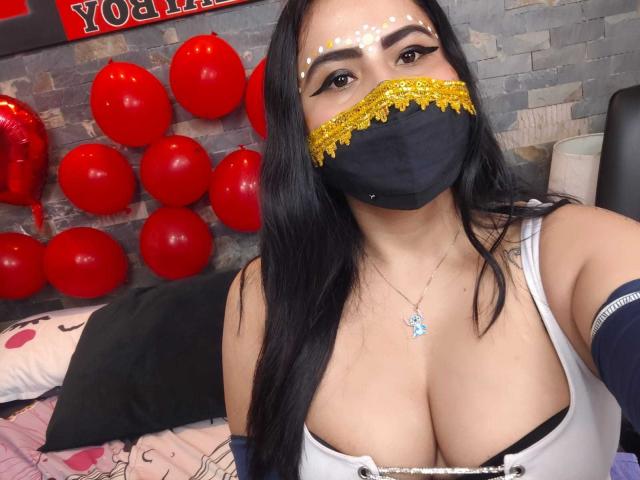 DaliaDubey with long black hair, wearing a black mask with gold sequins, white top, showing large boobs, gold necklace, and blue arm band, in a bedroom.