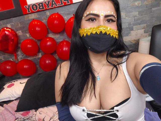 Dalia Dubey, met zwart haar, draagt een goud- en zwart masker, een witte top met een strik bij de hals, waardoor haar grote borsten zichtbaar zijn, en zit op een bed met rode ballonnen..