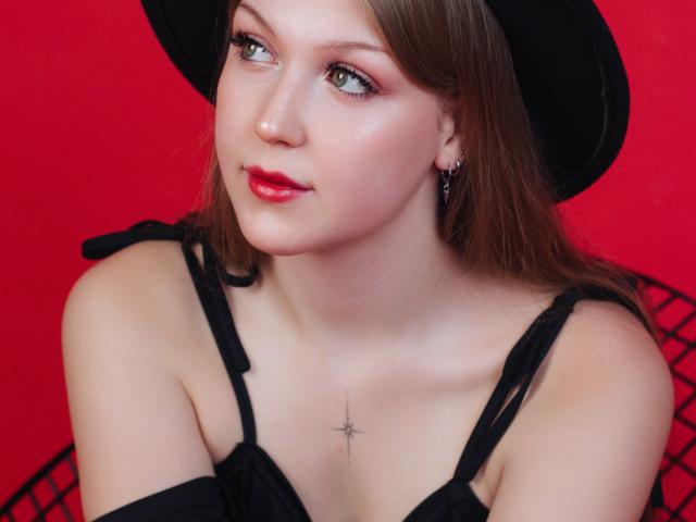 StellaLane with pale skin, red lipstick, and a black hat, showing off her tits and a small star tattoo on her chest, against a bright red background.