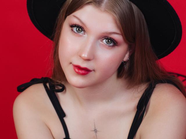 StellaLane with pale skin, green eyes, red lipstick, black hat, black straps, small cross tattoo below clavicle, brown hair, subtle makeup, smiling slightly, red background.