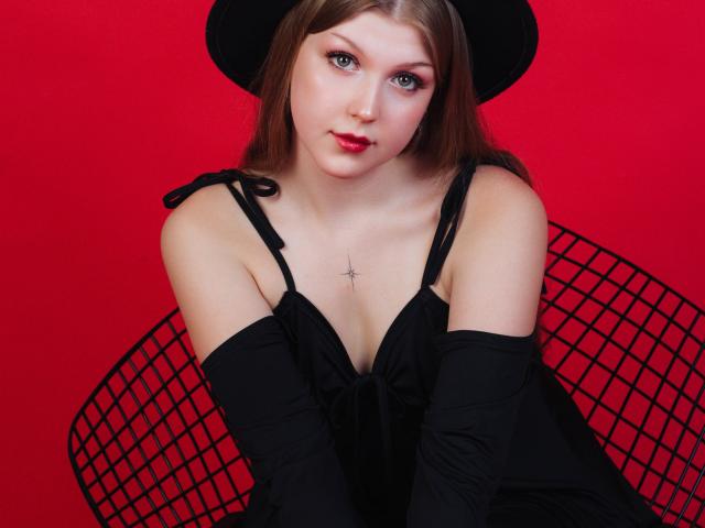 StellaLane has pale skin, blue eyes, and red lipstick, wearing a black dress with straps, long black gloves, and a black hat, sitting on a black wire chair.