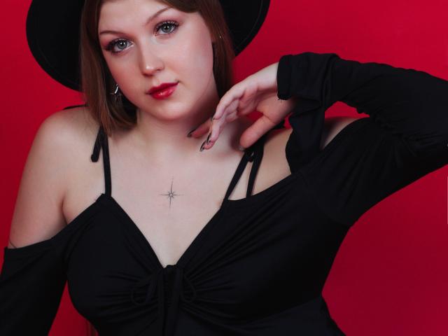 StellaLane, com pele clara, cabelo castanho e batom vermelho, vestindo um vestido preto e um chapéu, tem a mão perto do pescoço, mostrando uma tatuagem de estrela no peito..