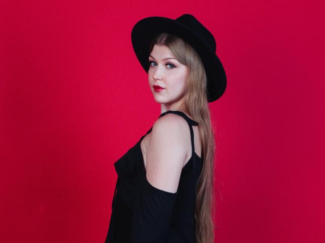 StellaLane with long blonde hair, black hat, and off-shoulder black dress, looking over shoulder, red background, red lips, pale skin, and subtle makeup. Boobs slightly visible.