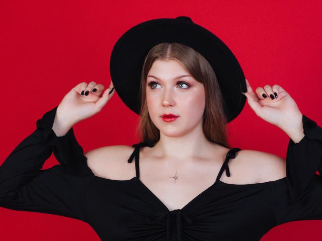 StellaLane with pale skin, red lipstick, and black nails, wearing a black off-shoulder top and hat, holding it, cross necklace, and looking to the side against a red background.