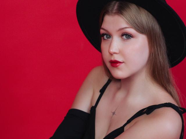 StellaLane tiene la piel pálida, lápiz labial rojo y ojos verdes. Lleva un sombrero negro y un top sin tirantes, mostrando un tatuaje de estrella en su pecho, con sus senos ligeramente expuestos..