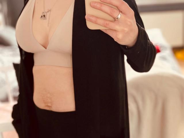 Il busto di Redhead88-hot in un selfie, che mostra la sua pelle chiara, piccoli seni in un reggiseno beige, un cardigan nero e un anello, mentre tiene un telefono, con una parte visibile dell'area pubica..