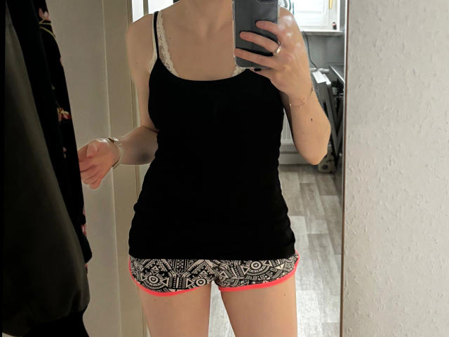 Redhead88-hot in black tank top with white lace trim, white and black patterned shorts with red trim, holding phone in mirror selfie. Her tits are small, her ass is not visible.