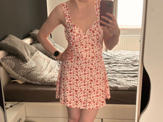 Redhead88, attraktiv in einem roten, blumigen Kleid, posiert in einem Spiegel-Selfie und zeigt ihre Brüste und einen kleinen Hintern. Sie trägt eine schwarze Armbanduhr und hält ein rosa Handy..