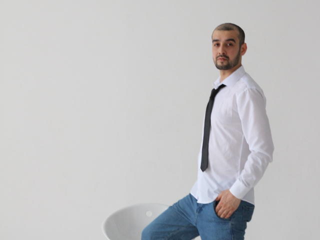 StormMigel with a clean-shaven face, short brown hair, white shirt, black tie, blue jeans, hand in pocket, standing next to a white chair, looking serious.