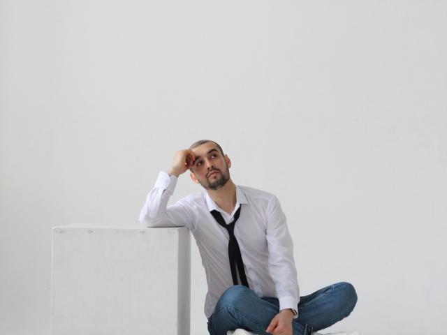 StormMigel sits with legs crossed, wearing a white shirt and black tie, hand on head, looking bored. He's got a beard, and his jeans are slightly wrinkled.