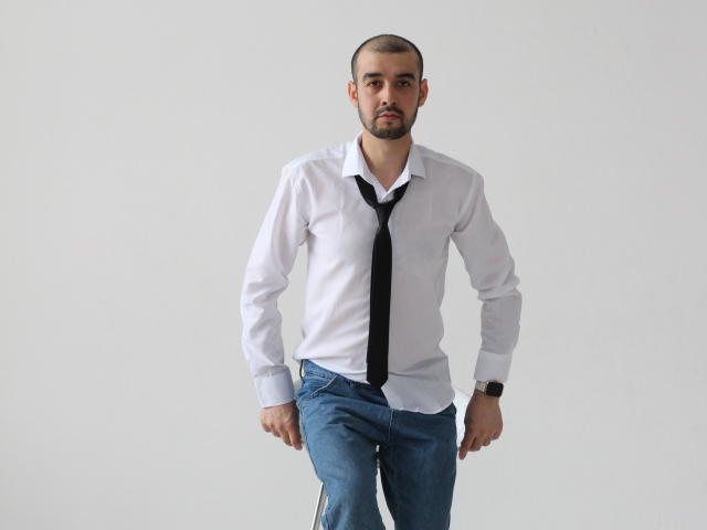 StormMigel with a trimmed beard, white shirt, black tie, and blue jeans, standing with a serious expression, wearing a black watch on his left wrist.
