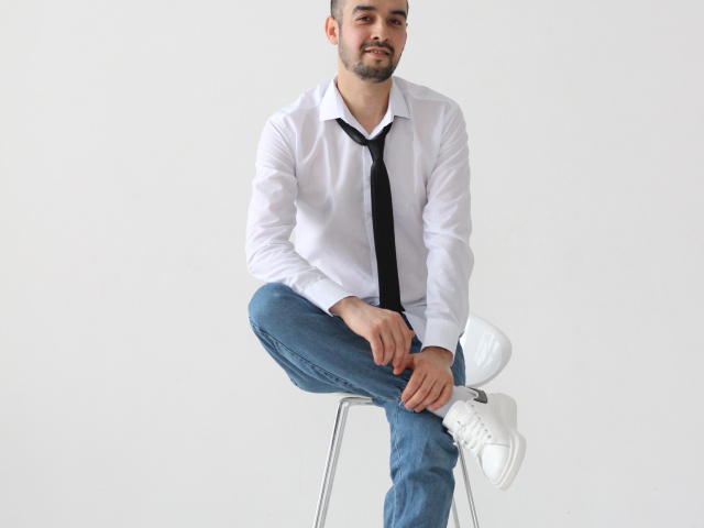 StormMigel sits on a stool, wearing a white shirt, black tie, blue jeans, and white sneakers. He has a short beard, a slight smile, and his legs are crossed.