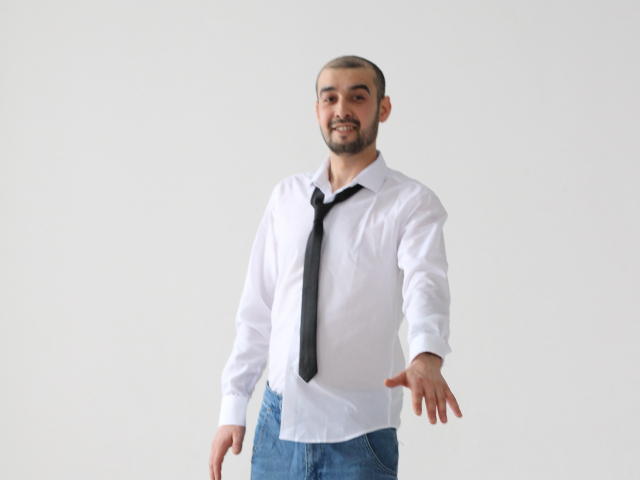 StormMigel, a dude with a trimmed beard, wearing a white shirt and black tie, standing with a relaxed smile, right arm out, hand open, and blue jeans.