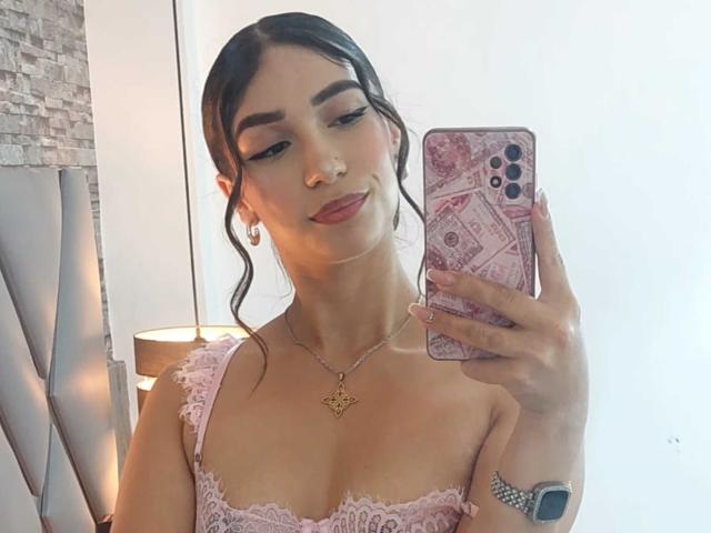 TanniaMoon con cabello oscuro en un moño suelto, blusa de encaje rosa, lengua afuera, sosteniendo un teléfono con funda rosa, collar dorado, aretes y reloj, mostrando sus senos..