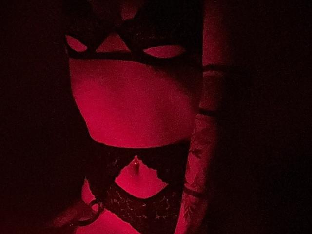 MiiS wearing black lace lingerie with cut-out eyes and a ringed navel, illuminated by red lighting, showing her bare midriff and lower chest.