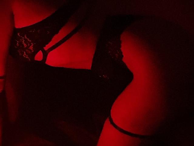 MiiS' black lace bra, revealing her boobs, with red lighting. Her right thigh, with a black strap, is in the foreground. The lace has a shiny, textured pattern.