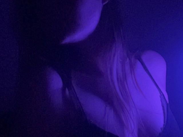 MiiS with visible tits, wearing a black strap, under purple light, showing her upper chest and part of her face, with long blonde hair.