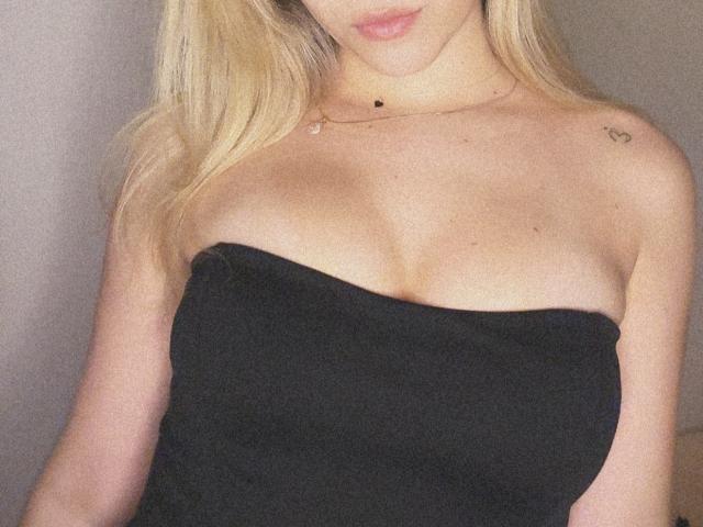 A SugarrX tem cabelo loiro, veste um top preto sem alças que realça seus seios grandes, com uma pequena tatuagem de coração no ombro direito e um colar delicado..
