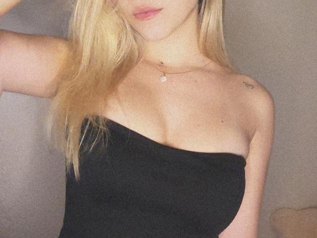 SugarrX with blonde hair, black strapless top, showing tits, small tattoo on right shoulder, necklace, pink lips, slightly blurred, soft lighting, gentle focus.