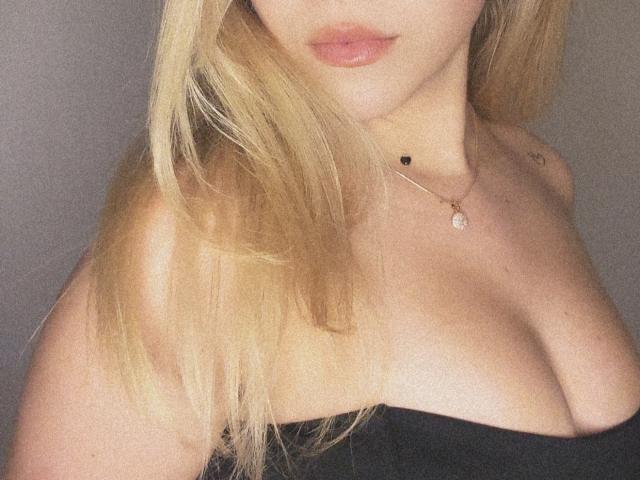SugarrX with blonde hair, wearing a black strapless top, showing her cleavage and a small black heart necklace with a gem. Soft, pinkish lips.