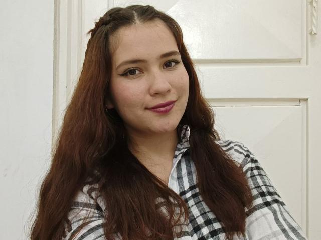 CelesteDash with long, wavy brown hair, wearing a black-and-white plaid shirt, smiling with pink lipstick, has cute, small tits, and a light skin tone.