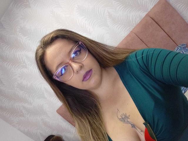 CleareRems with glasses, purple lipstick, green top, red bra, showing tits with a tattoo under her cleavage, long brown hair, looking at the camera.