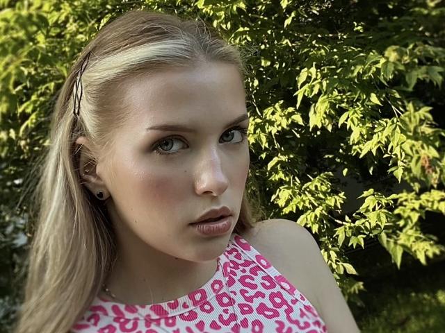FantsuyEva with blonde hair, green eyes, and pink leopard-print top, standing in front of green leafy background, looking straight at camera with slightly parted pink lips.
