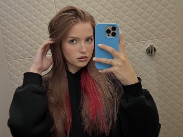 FantsuyEva with long brown and red hair, black top, holding blue phone, taking mirror selfie, right hand in hair, neutral expression, in a bathroom.