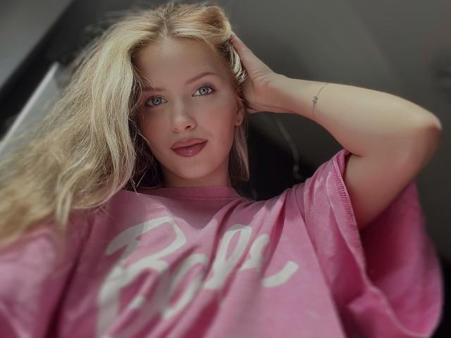FantsuyEva with blonde hair, blue eyes, and pink shirt, hand in hair, slight smile, pink lips, tattoo on wrist, soft light, casual, relaxed, sexy, cute.