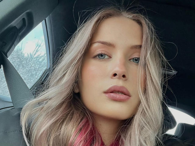 FantsuyEva's face in a car, with blonde hair, blue eyes, and pink lips, wearing a red scarf, looking slightly to the side, with a neutral expression.