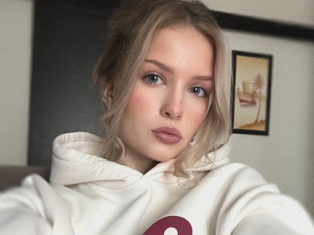 FantsuyEva has blonde hair, blue eyes, and pink lips, wearing a white hoodie. She's looking at the camera with a neutral expression. Her tits are small and she's cute.