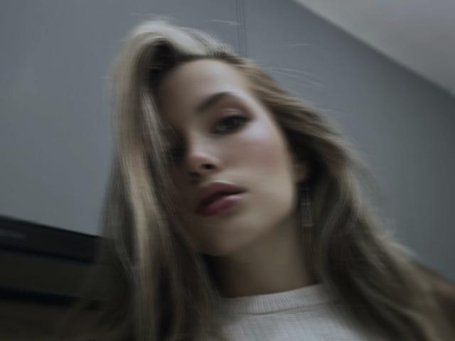 FantsuyEva has blonde hair covering one eye, full pink lips, and light skin. She's wearing a white sweater with her tits slightly visible. Blurred, low-angle shot.