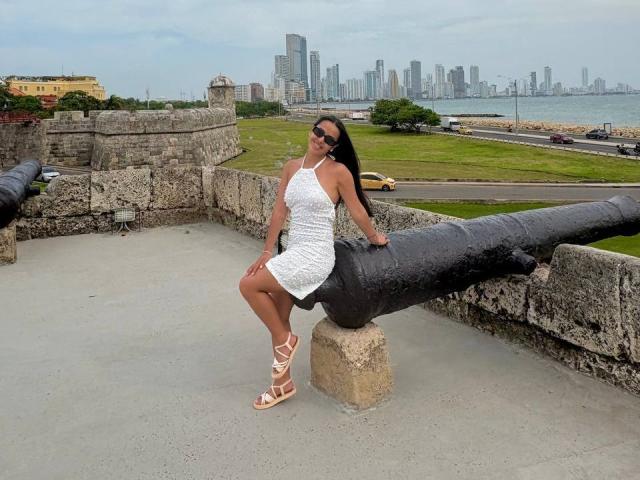 SamanthaRosses sitting on a large black cannon, wearing a white lace dress, strappy sandals, and sunglasses, with a city skyline and waterfront in the background.