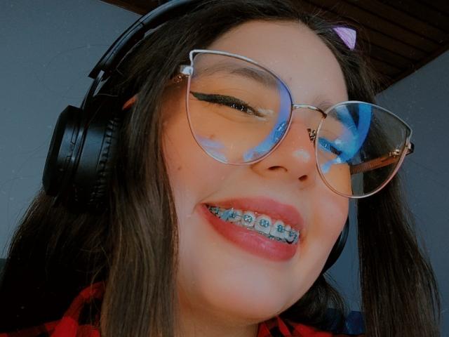 PaolaLyon with black headphones, glasses, and blue reflection in eyes, smiling with metal braces, wearing red plaid shirt, brown hair, purple hairclip, wooden ceiling above.