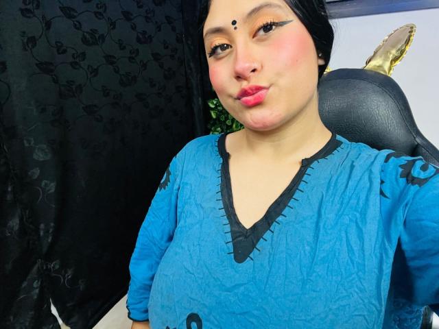 KalpanaAisha with pink lips, black headscarf, blue dress, black eyeliner, nose ring, and dark skin, pouting slightly, showing her chubby tits. Black curtain and chair in background.