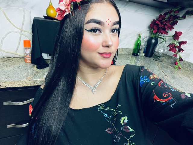 AnaShalimarr, com longos cabelos pretos, uma flor vermelha no cabelo, um vestido preto com bordados coloridos, batom vermelho e um colar de diamantes, está em uma cozinha, olhando para a câmera..